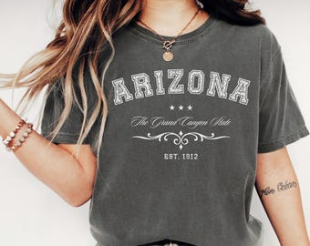 May include: A dark gray t-shirt with the word "ARIZONA" in white, vintage-style lettering. Below the word "ARIZONA" are three stars, the text "The Grand Canyon State", and the year "EST. 1912". The shirt is a solid color and has short sleeves.