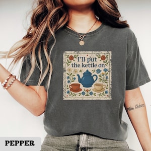 May include: A dark gray t-shirt featuring a vintage-style graphic with the text "I'll put the kettle on." The design includes a blue teapot, two teacups, and floral elements in a square frame. The word "PEPPER" is at the bottom.
