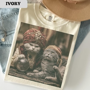 May include: Ivory-colored t-shirt featuring a detailed graphic of two cats dressed in regal attire, one holding a crystal ball. The cats wear turbans and ornate headwear. The shirt has a crew neck and a tag that reads "Comfort Colors".