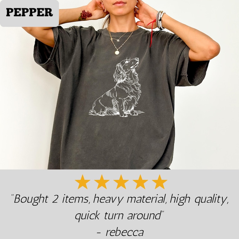 May include: Dark gray t-shirt featuring a white line drawing of a long-haired dachshund. The shirt has short sleeves and a relaxed fit. The word "PEPPER" is in a white box in the upper left corner.
