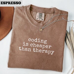 May include: Espresso-colored t-shirt with the text "coding is cheaper than therapy" in white. The shirt has a crew neck and short sleeves. A silver necklace with a small pendant is draped over the shirt.