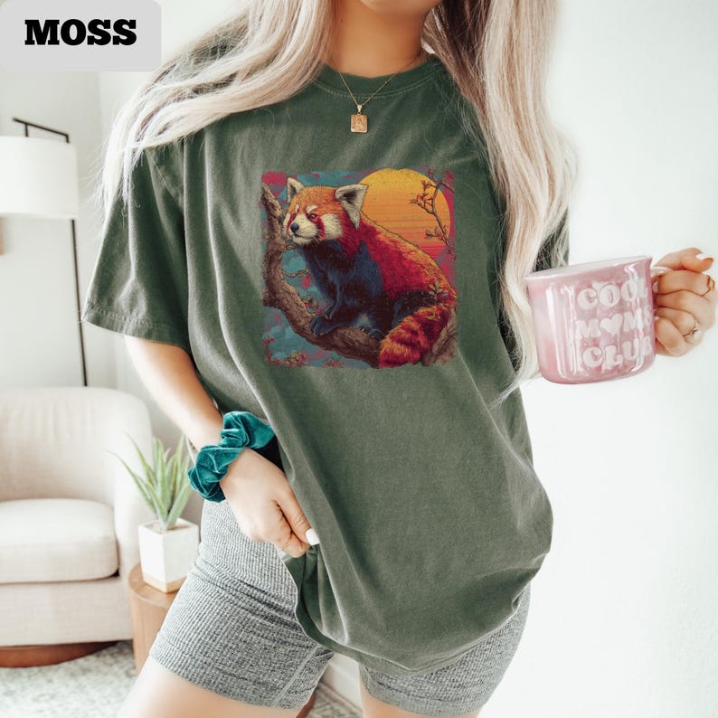 Puede incluir: Camiseta verde musgo con un dise&ntilde;o de panda rojo sobre un fondo de puesta de sol. El panda rojo est&aacute; posado en una rama. La camiseta es de corte holgado. El texto "MOSS" est&aacute; en la esquina superior izquierda.