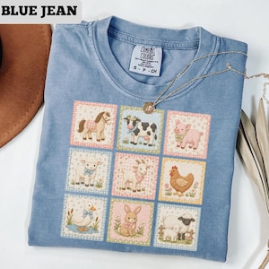 May include: Blue jean colored t-shirt with a patchwork design featuring farm animals. The shirt has a crew neck and short sleeves. The text "BLUE JEAN" is at the top left. A gold necklace is draped across the shirt.