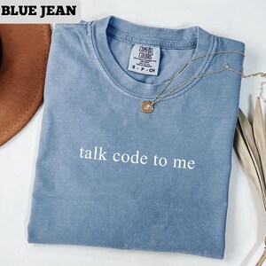 May include: A blue jean colored t-shirt with the text "talk code to me" in white. The shirt has a crew neck and short sleeves. A gold necklace with a pendant is draped across the shirt.