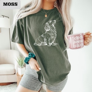 May include: Moss green t-shirt with a white illustration of a long-haired dachshund. The shirt has a relaxed fit. The word "MOSS" is in the upper left corner. A pink mug with the words "Cool Moms Club" is held in the hand.
