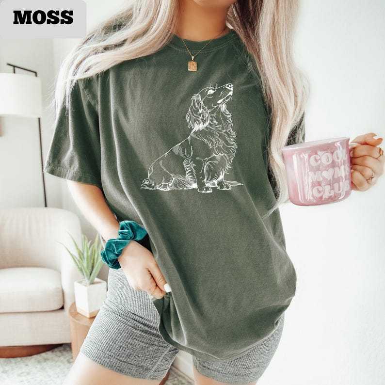 May include: Moss green t-shirt with a white illustration of a long-haired dachshund. The shirt has a relaxed fit. The word "MOSS" is in the upper left corner. A pink mug with the words "Cool Moms Club" is held in the hand.