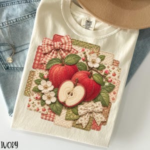 May include: Ivory-colored t-shirt featuring a detailed apple design. The design includes red apples, green leaves, white flowers, and decorative bows. The word "IVORY" is printed at the bottom. The shirt is folded on top of blue jeans.