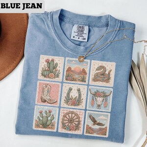 May include: Blue jean colored t-shirt with a grid of nine southwestern-themed images. The images include a cactus, desert landscape, snake, cowboy boot, horseshoe, steer skull, wagon wheel, and an eagle. The shirt has the text "BLUE JEAN" at the top.