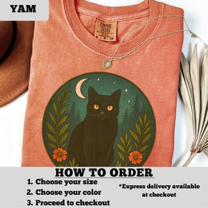 May include: A coral-colored t-shirt featuring a circular graphic of a black cat with yellow eyes, a crescent moon, and a forest scene. The shirt has a Comfort Colors tag and a gold necklace. Text on the shirt reads "HOW TO ORDER".