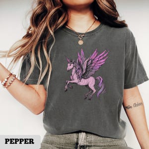 May include: A dark gray t-shirt featuring a pink unicorn with purple wings. The unicorn is depicted in a running pose. The word "PEPPER" is printed in white at the bottom of the shirt.