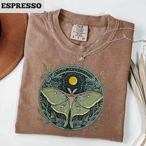 May include: Espresso-colored t-shirt featuring a large green moth design within a circular frame. The design includes a yellow moon and stars. The shirt has a crew neck and short sleeves. A silver necklace with a pendant is draped across the shirt.