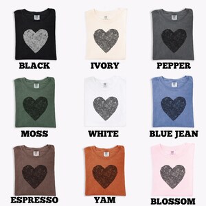 K&ouml;nnte beinhalten: Neun verschiedenfarbige T-Shirts, jedes mit einem gro&szlig;en Herz-Grafik. Farben sind Schwarz, Elfenbein, Pfeffer, Moos, Wei&szlig;, Blue Jean, Espresso, Yam und Blossom. Jedes Shirt hat den Farbnamen darunter gedruckt.