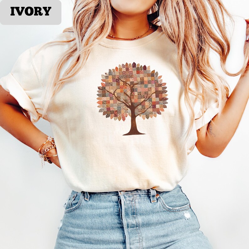 May include: Ivory-colored t-shirt featuring a tree design. The tree has a brown trunk and branches, with a patchwork quilt-style canopy in various colors. The word "IVORY" is printed in a rectangular box at the top left.