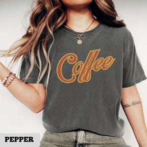 May include: A gray t-shirt with the word "Coffee" in a retro, orange and gold font. The shirt is a short-sleeved, crew neck style. The word "Pepper" is at the bottom of the image.
