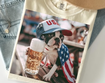 May include: An ivory t-shirt featuring a graphic of a Boston Terrier wearing a red, white, and blue USA baseball cap and a flag-patterned cape, holding a glass mug of beer. The shirt has a crew neckline and a Comfort Colors brand label inside.