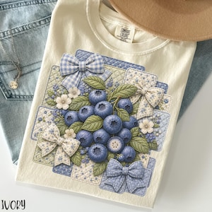 May include: Ivory-colored t-shirt featuring a detailed illustration of blueberries, green leaves, and decorative bows. The design includes floral and patchwork elements. The shirt is paired with blue jeans and a necklace.