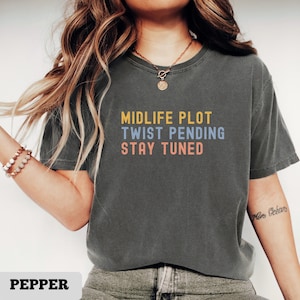 May include: A pepper-colored t-shirt with the text "MIDLIFE PLOT TWIST PENDING STAY TUNED" in yellow, blue, and orange. The shirt has a crew neck and short sleeves.