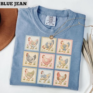 May include: Blue t-shirt with a square chicken pattern. The shirt has the text "BLUE JEAN" at the top left. A gold necklace with a pendant is draped across the shirt. The shirt is labeled "COMFORT COLORS" on the tag.