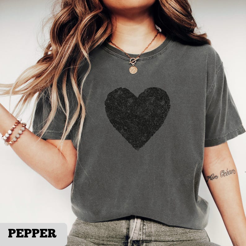 K&ouml;nnte beinhalten: Graues T-Shirt mit einem gro&szlig;en schwarzen Herzdesign. Das Shirt hat einen Rundhalsausschnitt und kurze &Auml;rmel. Das Wort "PEPPER" ist unten im Bild aufgedruckt.