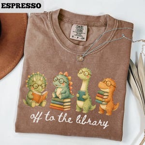 May include: Espresso-colored t-shirt featuring a graphic of four cartoon dinosaurs wearing glasses and holding books. The text "off to the library" is printed below the dinosaurs. A silver necklace with a pendant is draped on the shirt.