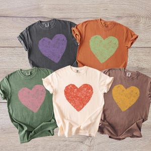 K&ouml;nnte beinhalten: F&uuml;nf T-Shirts in verschiedenen Farben, jedes mit einem verblassten Herzmotiv. Farben sind Anthrazitgrau mit lila Herz, Orange mit gr&uuml;nem Herz, Olivgr&uuml;n mit rosa Herz, Creme mit rotem Herz und Braun mit gelbem Herz.