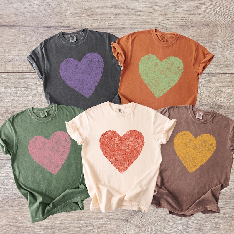 K&ouml;nnte beinhalten: F&uuml;nf T-Shirts in verschiedenen Farben, jedes mit einem verblassten Herzmotiv. Farben sind Anthrazitgrau mit lila Herz, Orange mit gr&uuml;nem Herz, Olivgr&uuml;n mit rosa Herz, Creme mit rotem Herz und Braun mit gelbem Herz.