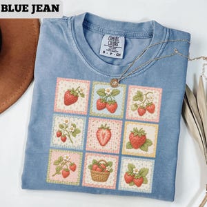 May include: Blue jean Comfort Colors t-shirt with a strawberry patchwork design. The shirt features a crew neck and short sleeves. A gold necklace with a pendant is draped across the shirt. The text "BLUE JEAN" is at the top.
