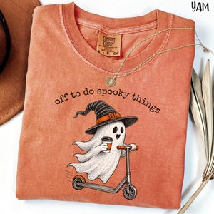 May include: A terracotta-colored crew neck t-shirt featuring a graphic of a ghost wearing a witch hat and riding a scooter while holding a coffee cup. The text "off to do spooky things" is printed above the ghost in a black, rounded font.