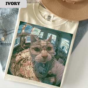May include: Ivory-colored t-shirt featuring a cat wearing glasses and a blue uniform, with a control room background. The shirt has a Comfort Colors tag. The word "IVORY" is printed in the upper left corner.