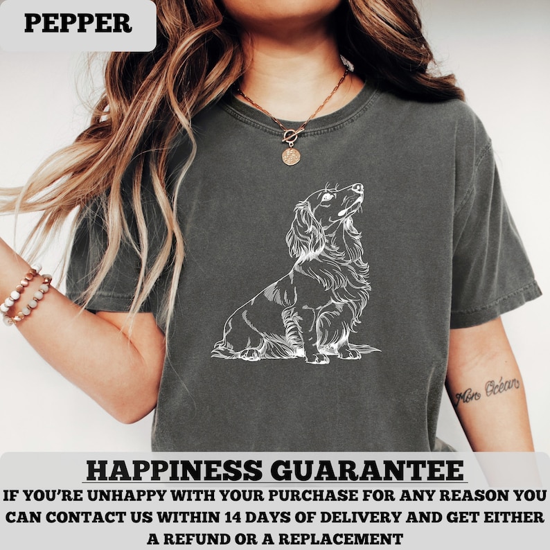 May include: Dark gray t-shirt featuring a white illustration of a long-haired dachshund looking upwards. The word "PEPPER" is in a white box in the upper left corner. The text "HAPPINESS GUARANTEE" is at the bottom.