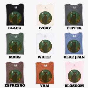 May include: Nine different colored t-shirts are displayed, each with a graphic of a black cat inside a circle with a moon and flowers. The shirt colors include black, ivory, pepper, moss, white, blue jean, espresso, yam, and blossom.