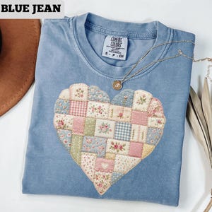 May include: Blue jean colored t-shirt with a patchwork heart design. The heart is made of various floral and patterned fabrics in pastel colors. A gold necklace with a pendant is draped on the shirt. The text "BLUE JEAN" is at the top.