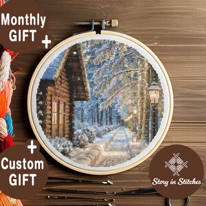 Könnte beinhalten: Ein Kreuzstich-Stickrahmen mit einer Winterszene mit einer Hütte und einem schneebedeckten Weg. Der Holzrahmen hält die detaillierte Handarbeit, mit dem Text "Story in Stitches" und "Monthly GIFT + Custom GIFT" sichtbar.