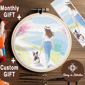May include: A cross-stitch embroidery hoop featuring a woman walking a dog on a path, with a pink tree and blue sky background. The text "Monthly GIFT +" and "+ Custom GIFT" are visible. A bookmark with a similar design is also present.