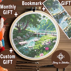 May include: A cross-stitch landscape scene in a wooden embroidery hoop, depicting a serene river scene with a bridge, trees, and flowers. A matching bookmark and the words "Monthly Gift" and "Custom Gift" are also visible.