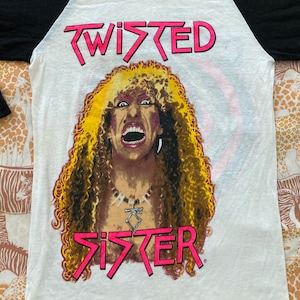 May include: A white and black baseball tee shirt with a colorful image of a man with long blonde hair and the words "Twisted Sister" printed on the front.