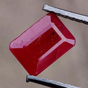 Peut inclure: Une pierre précieuse rubis rouge à facettes rectangulaires tenue par une pince en métal. La pierre présente une teinte cramoisie profonde avec des inclusions internes visibles et des bords nets le long de sa surface taillée.