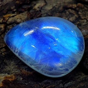 May include: A polished, oval-shaped gemstone with a mesmerizing blue iridescence. The stone's surface reflects light, creating a luminous effect. The gem is set against a dark, textured background, highlighting its vibrant color and unique patterns.