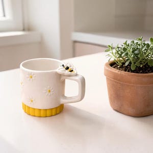 May include: A white ceramic mug with daisy accents and a yellow base. The handle features a bee design. A small potted plant with green foliage sits beside the mug on a white surface.