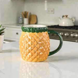 Peut inclure: Une tasse en céramique en forme d'ananas, avec un corps jaune et des détails verts. La tasse a une surface texturée et une anse verte. L'intérieur est blanc. Une petite plante succulente dans un pot blanc est à gauche.