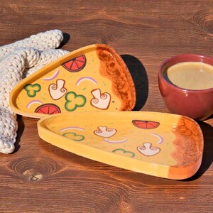 May include: Two pizza-shaped serving platters with yellow bases and colourful toppings, including mushrooms, green peppers, and tomato slices. A maroon coffee cup with coffee sits next to the platters. A white knitted blanket is in the background.
