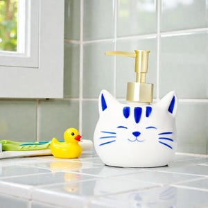 Op de afbeelding: Een witte keramische zeepdispenser in de vorm van een kattenkop, met blauwe accenten en een gouden pomp. Een gele badeend en een groen-witte tandenborstel zijn ook zichtbaar op een betegeld oppervlak.