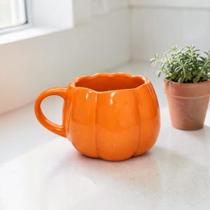 Peut inclure: Une tasse en céramique orange en forme de citrouille avec une anse. La tasse est à côté d'une petite plante en pot avec des feuilles vertes. La tasse est sur une surface blanche.