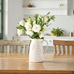 Könnte beinhalten: Eine cremefarbene Keramikkrug mit floralem Design hält einen Strauß weißer Blumen und grüner Stiele. Der Krug steht auf einem Holztisch, im Hintergrund sind Holzstühle zu sehen. Eine Uhr und ein Schrank sind ebenfalls sichtbar.