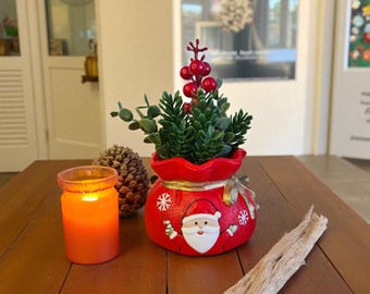 May include: A festive Christmas-themed arrangement featuring a red Santa Claus-decorated pot filled with greenery and red berries. A lit orange candle in a glass jar and a pinecone sit nearby on a wooden surface. A piece of driftwood is also present.