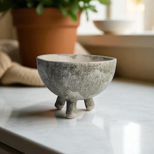 Könnte beinhalten: Eine kleine, handgefertigte Keramikschale mit gesprenkelter grau-weißer Oberfläche. Die Schale hat eine abgerundete Form und steht auf drei kurzen, stabilen Beinen. Der Innenraum ist heller, cremefarben. Diese dekorative Schale eignet sich für kleine Pflanzen.