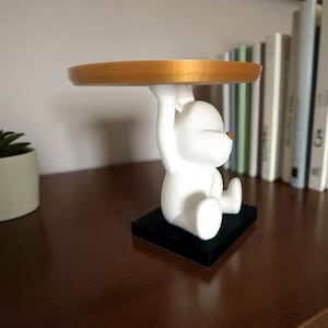 以下が含まれることがあります： 金色のトレイを持った白いクマの置物。クマは黒い台座に座っています。この置物は、小さなアイテムを置いたり、ディスプレイとして使用したりするための装飾品です。