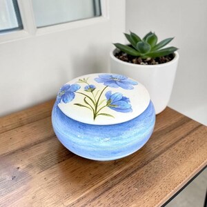 May include: A round ceramic lidded jar with a light blue painted base and a white lid featuring a hand-painted design of blue flowers with green stems and leaves. The jar has a smooth, glossy finish and a rounded shape, sitting as a decorative container.