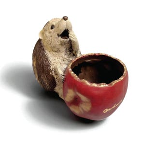 Puede incluir: Una caprichosa figura de cerámica de erizo con expresión de sorpresa, sosteniendo un cuenco en forma de manzana roja. La manzana tiene un borde dorado y una pequeña inscripción. El erizo tiene pelaje marrón y beige.