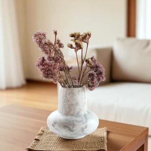 Puede incluir: Un jarrón blanco y gris con vetas de mármol que contiene flores secas moradas y tostadas. El jarrón tiene una base redondeada y un cuello más estrecho. Se encuentra sobre una mesa de madera con un tapete tejido.